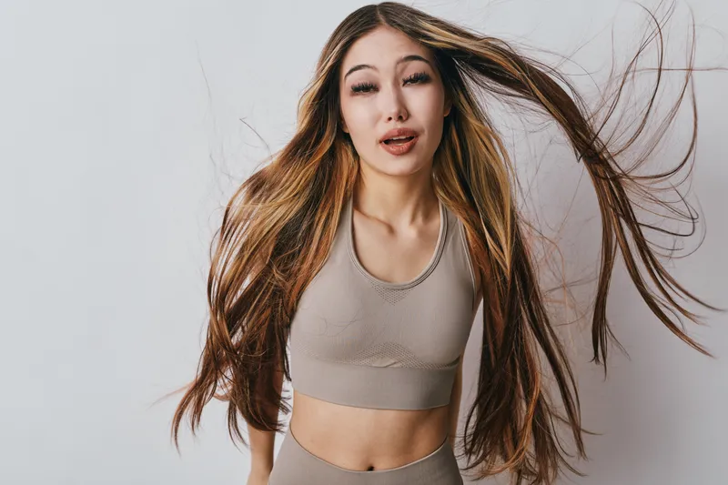 Young woman with long hair in motion wearing beige sportswear posing on light background. Dynamic hair movement, female fitness model with slim figure and active lifestyle concept.