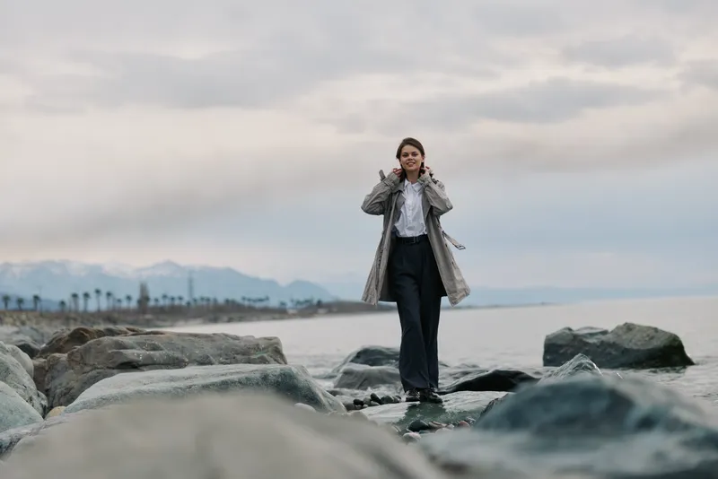Young woman in casual clothes and jacket walks on rocks near sea coast on cloudy day. Concept of outdoor lifestyle, nature exploration, and peaceful scenery with mountains and water background.