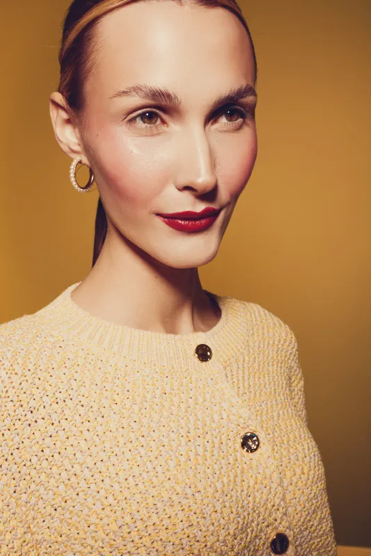 Close-up portrait of a confident woman with red lipstick and hoop earrings wearing a yellow knitted sweater against a warm yellow background. Concept of fashion, beauty, and elegance.