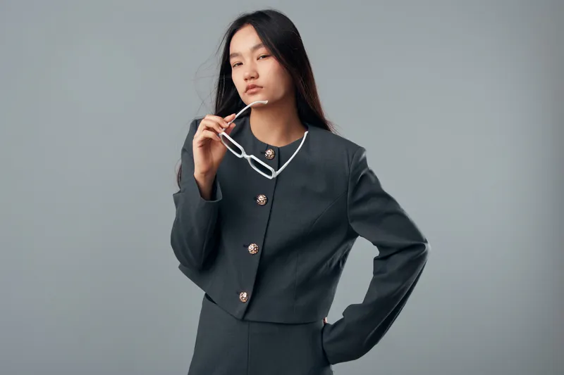 Young woman with long black hair wearing dark suit holding white eyeglasses and looking serious on plain background. Female model posing with hand on hip for fashion portrait.