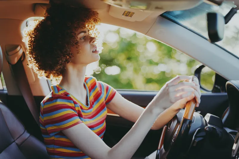 Woman driving a car with curly hair, striped shirt, sunlight through trees, cheerful expression, urban road scene, vibrant retro hues, natural shadows, relaxed posture, candid lifestyle moment,