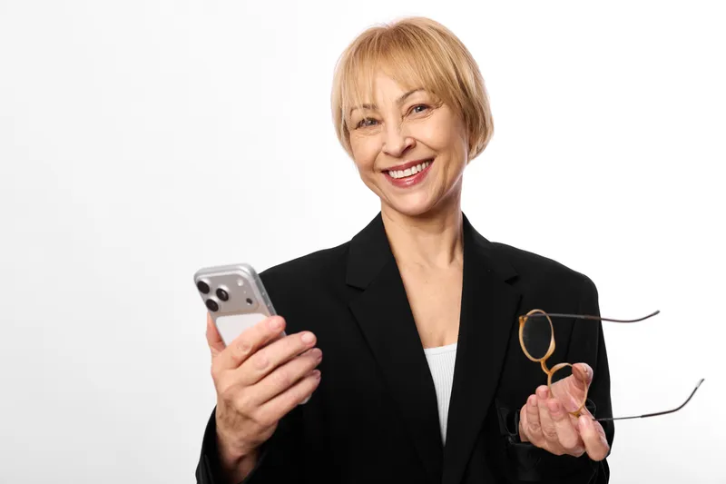 Woman businesswoman smartphone glasses smile suit isolated white background, smiling senior woman in black blazer holds smartphone and eyeglasses, studio portrait on plain white