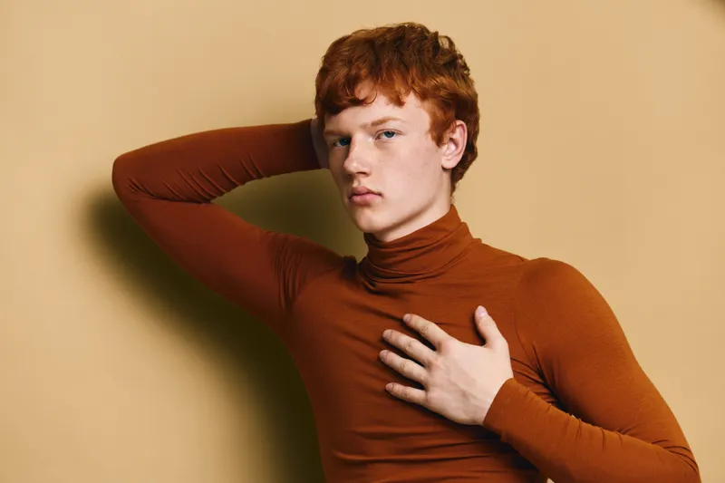 Young man with red hair posing in brown turtleneck sweater against beige background, portrait with serious expression and hand resting on chest and head for fashion or style concept.