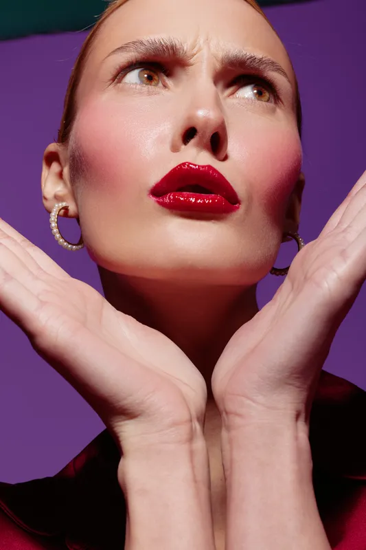 Close-up of a woman with glossy red lips and bold makeup posing with hands near face on purple background. Concept of beauty, fashion, and makeup artistry in studio lighting.