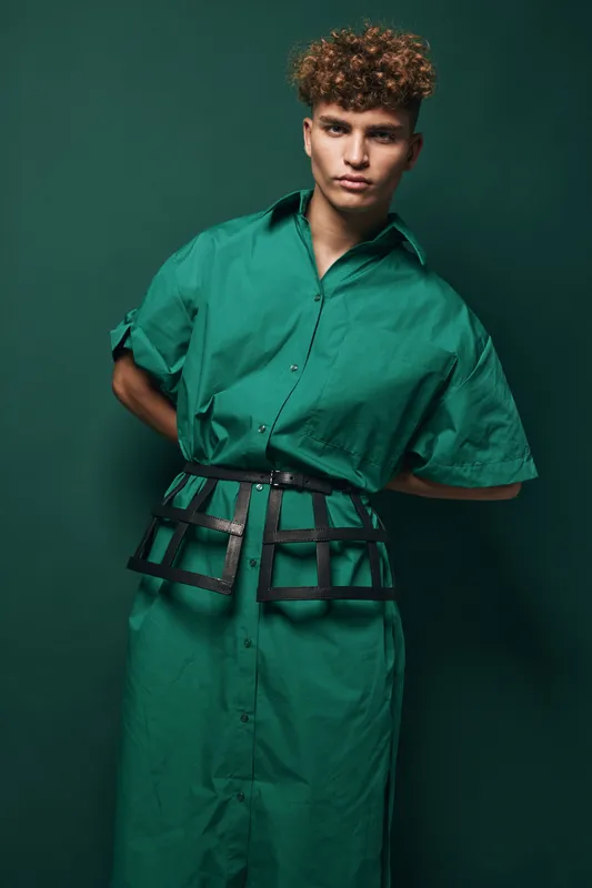 Young man with curly hair wearing stylish green dress with black statement belt posing indoors, contemporary fashion portrait with confident expression on dark green background.
