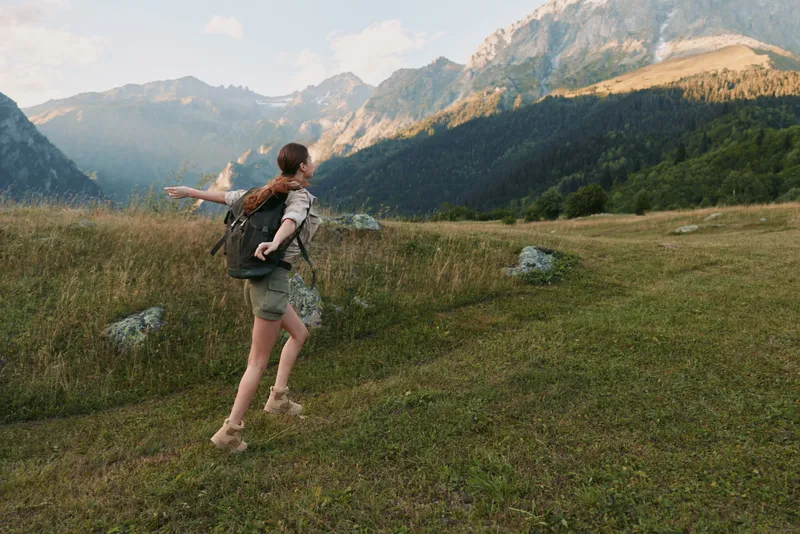 Young woman with a backpack runs across a grassy meadow toward distant mountains, capturing outdoor adventure, freedom, and a carefree travel lifestyle.