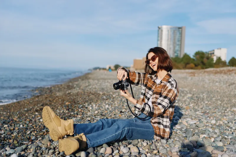 Casual photographer on a pebbled beach, kneeling with a camera, wearing a plaid shirt and boots, capturing coastal scenery and sunlight in a relaxed outdoor moment of photography