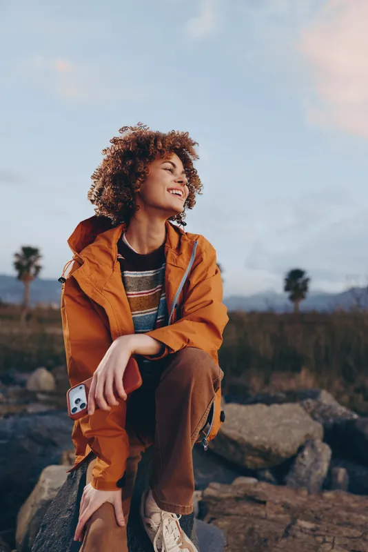 Smiling woman lifestyle outdoors during hiking on rocky trail, orange jacket, casual jeans, curly hair in wind, holding smartphone, scenic landscape with palms and mountains at dusk