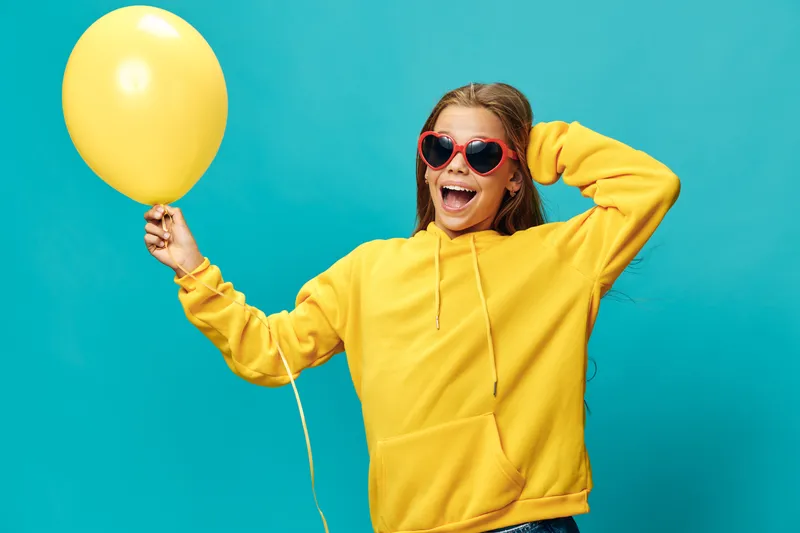 Girl in yellow hoodie and red heart sunglasses holding yellow balloon on blue background with joyful expression smiling for fun and celebration event or party moment.