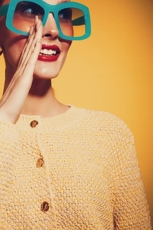 Smiling woman in large turquoise sunglasses and yellow knitted sweater poses against yellow background. Bright makeup and joyful expression create vibrant, fashionable portrait concept.