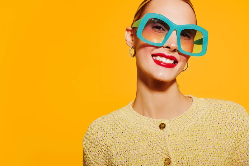 Smiling woman with bright red lipstick wearing large blue sunglasses and yellow knitted sweater on vibrant yellow background. Concept of fashion, summer style, and confident mood.