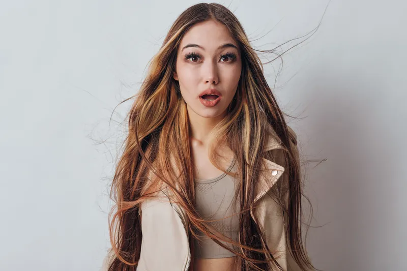 Woman with long hair and surprised expression looking at camera against white background. Casual outfit with jacket and natural makeup for fresh portrait.