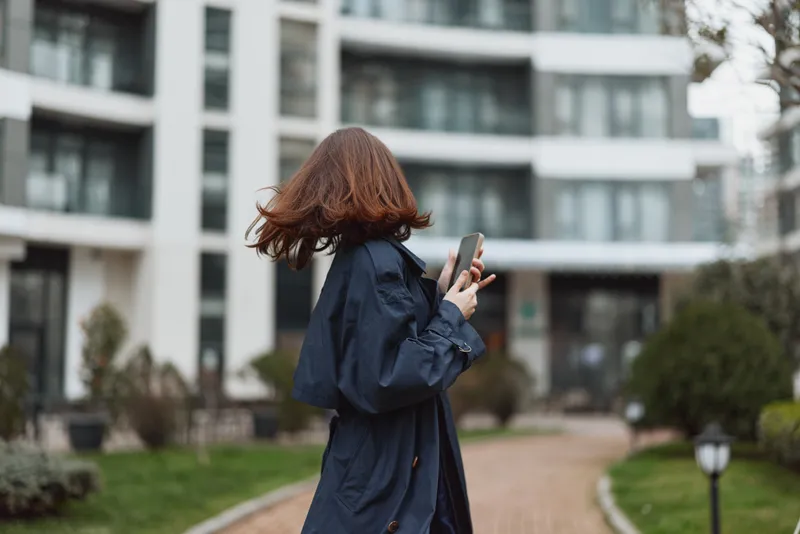 A young woman with red hair wearing a dark blue coat walks outdoors while looking at her smartphone in an urban residential area during daytime.