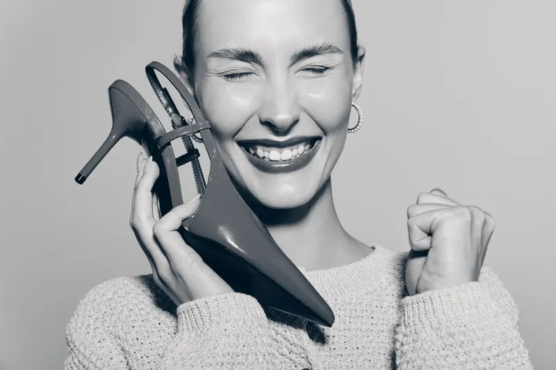 Happy caucasian woman with fair skin closes eyes and smiles while holding high heel shoes near face in black and white photo.