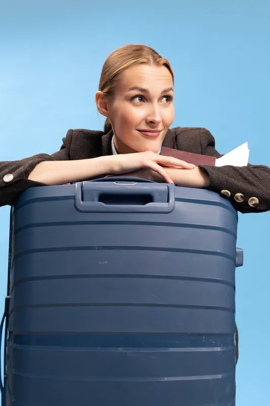 Young woman resting on large blue suitcase looks away smiling on light blue background. Concept for travel, vacation, luggage, and holiday planning.