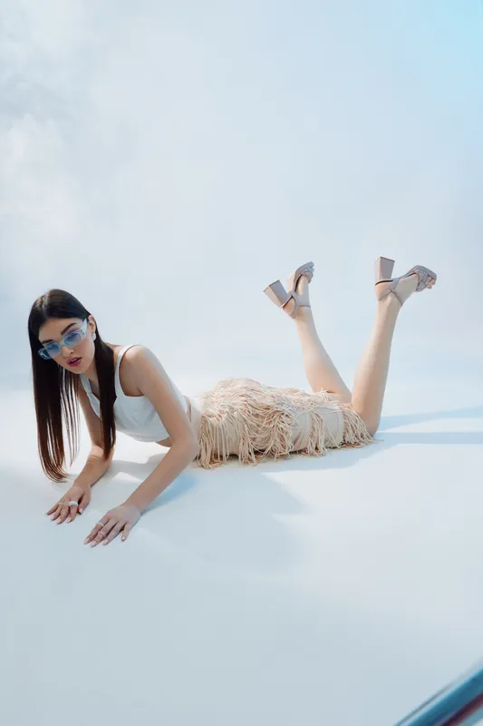 Young woman in stylish beige fringe skirt and white top lies on floor wearing sunglasses. Fashion concept with beauty, elegance, and modern style on light background.
