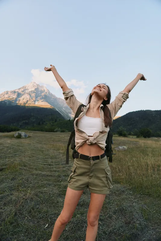 A woman outdoors raises her arms in triumph against a mountain backdrop, wearing casual hiking attire. The scene emphasizes lifestyle, freedom, nature, and adventure in a vibrant outdoor setting.