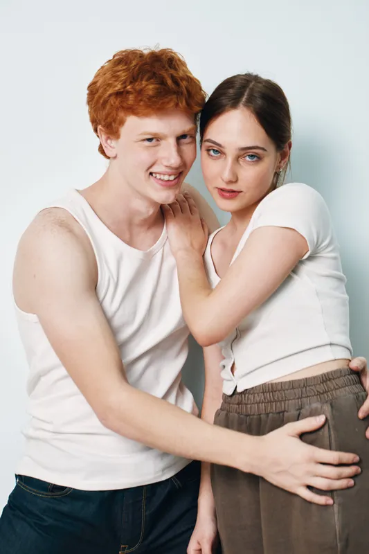 Young couple with relaxed expressions posing in casual clothes against white background close together with affectionate and confident body language indoors in studio light.