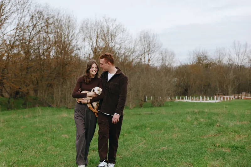 Young couple walks together in a grassy field holding a baby goat while enjoying a peaceful autumn day with bare trees and overcast sky in the background.