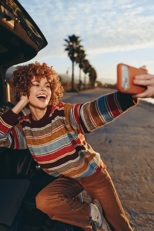 Happy woman taking selfie outdoors beside car on road, smiling with curly hair, holding phone up, warm sunset light, palm trees blurred background, casual lifestyle, relaxed pose and laugh