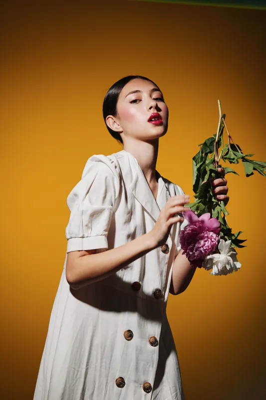 Fashion model with flawless makeup holds peony bouquets against bright orange background. White blazer dress and bold lips create creative beauty concept for style editorial.