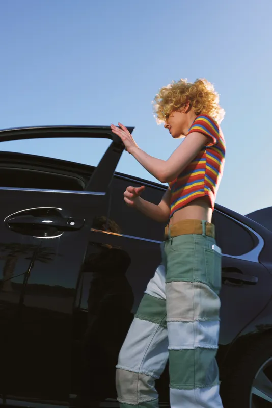 Young woman with curly blonde hair in a striped top and patchwork jeans opens a car door outdoors, showcasing casual street style and contemporary fashion on a sunny day