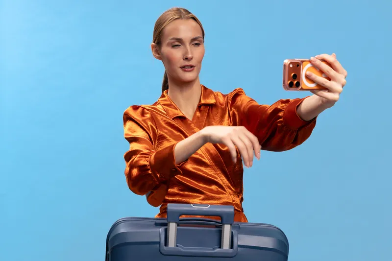 Young woman in orange blouse takes selfie with smartphone while standing behind blue suitcase on blue background. Concept of travel, vacation preparation, and modern technology use.