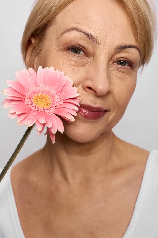 elderly woman beauty portrait in studio, holding pink gerbera flower by cheek, calm smile, visible wrinkles, skincare concept, natural glow for wellness
