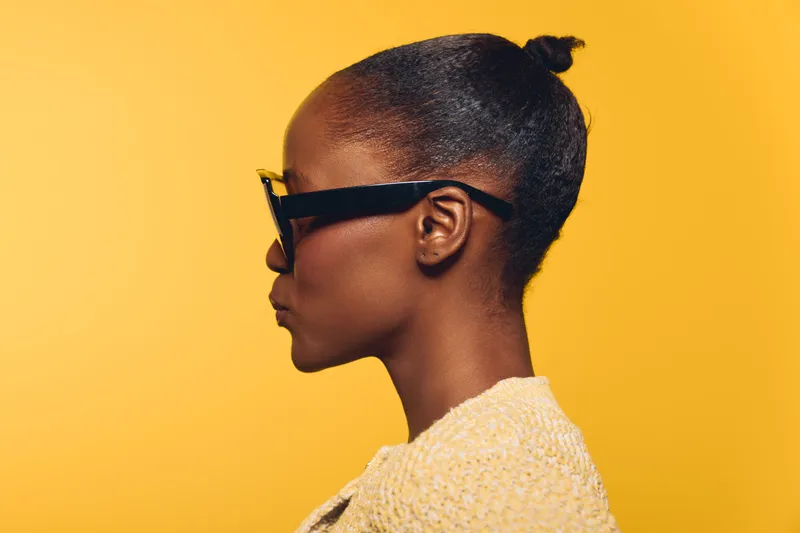 Profile of a young African woman with dark skin wearing black sunglasses and a beige knitted sweater on yellow background. Concept of fashion, style, and confident attitude.