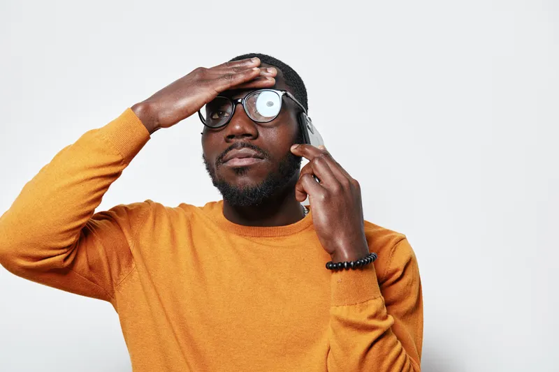 African man with dark skin wearing glasses and orange sweater talking on smartphone with worried expression and hand on forehead against white background.