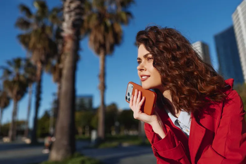 Young adult brunette woman in a red coat chats on her phone outdoors amid a sunny urban backdrop with palm trees in the distance, a bright sky and a candid fashion moment.