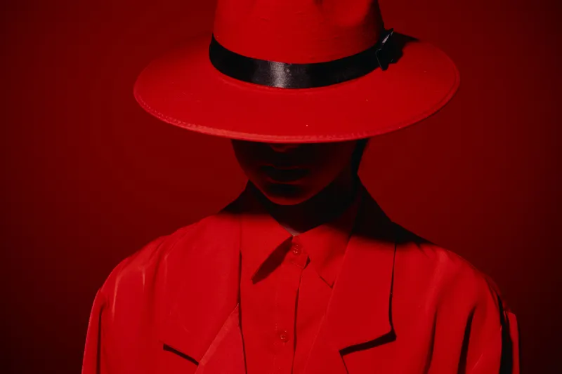 Fashion model in red hat and jacket poses against red background. The shadowed face adds mystery and creativity, capturing a bold beauty and fashion concept with dramatic lighting.