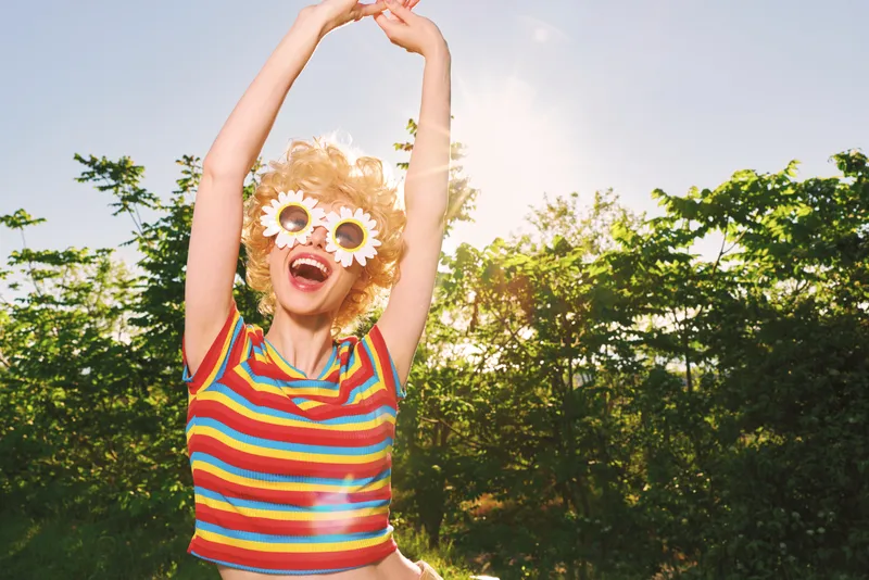 A joyful woman outdoors raises her arms in a sunny park, wearing a striped shirt and flower sunglasses. Retro film color tones emphasize carefree lifestyle and summer bliss.