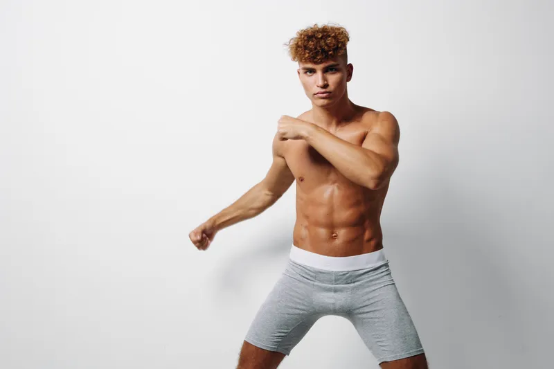 Young muscular man with curly hair posing shirtless in gray shorts against white background, showing fitness, strength, and confident expression in studio lighting.