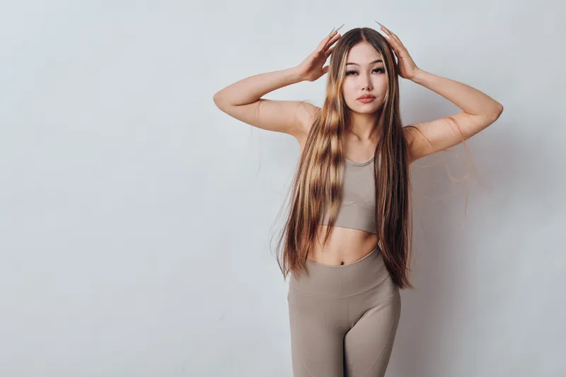 Young woman with long hair poses with hands on head wearing beige two-piece outfit in front of white wall. Beauty, casual style and confident facial expression indoors.