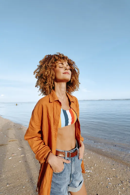 A young woman with curly hair stands on a sunny beach in a casual orange shirt over a striped bikini, eyes closed, embracing the warm breeze and carefree summer mood.