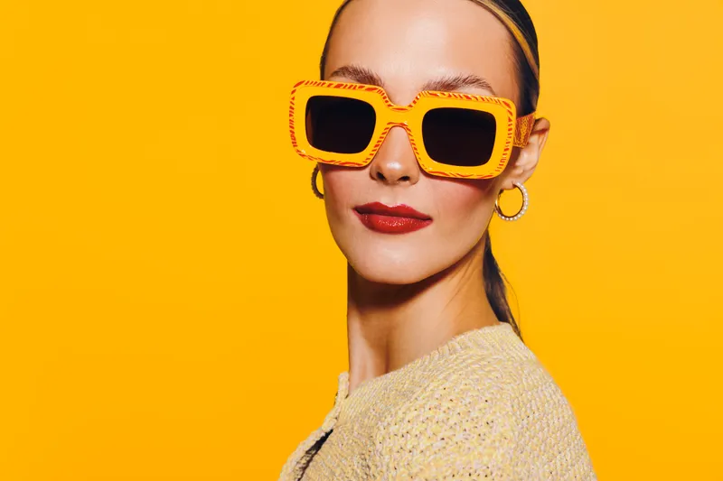 Stylish woman with slicked back hair wears large orange sunglasses and red lipstick against bright yellow background. Concept of fashion, summer style, and confident beauty portrait.