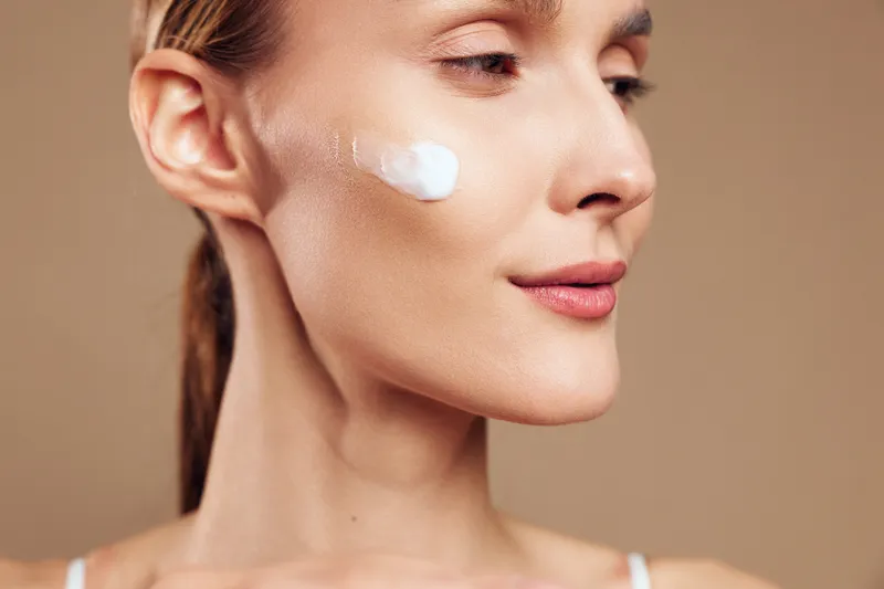 Young woman with brunette hair applies white face cream on her cheek while closing her eyes in a calm skincare routine against a soft brown background.