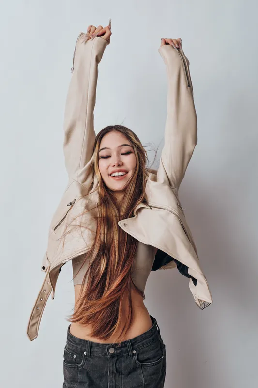 Young woman with long hair wearing casual jacket and jeans, smiling and raising arms up, isolated on white background in studio. Positive emotion, freedom, happiness and style.