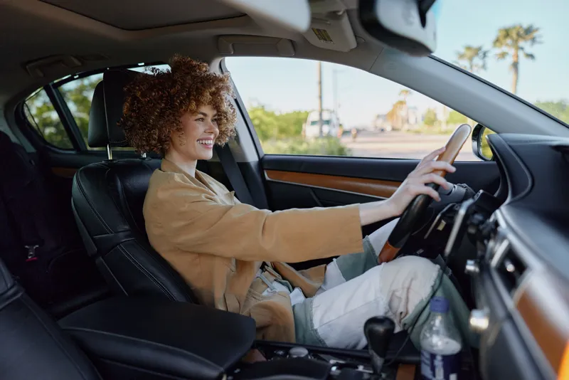 Woman drives a modern car with a bright smile, casual outfit, sunlit interior, and relaxed mood, showcasing everyday travel and independent style in a candid lifestyle photograph