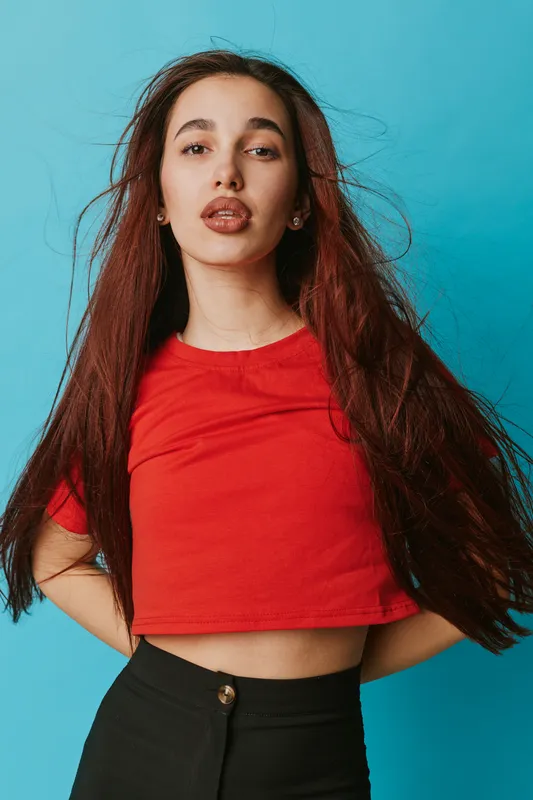 Young woman with long brown hair wearing red crop top and black pants posing confidently against blue background. Portrait with natural makeup and casual fashion style in studio setting.