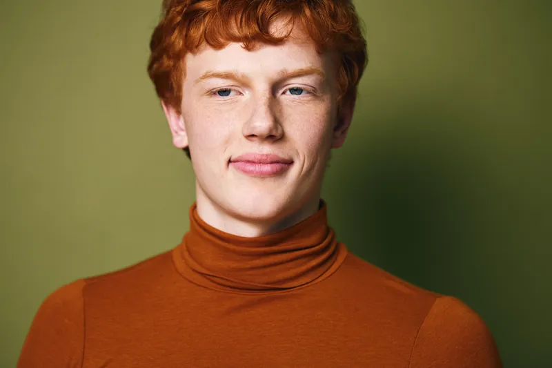 Portrait of redhead young man wearing high neck brown sweater with confident expression on green background, closeup of casual male model with fair skin and curly hair.