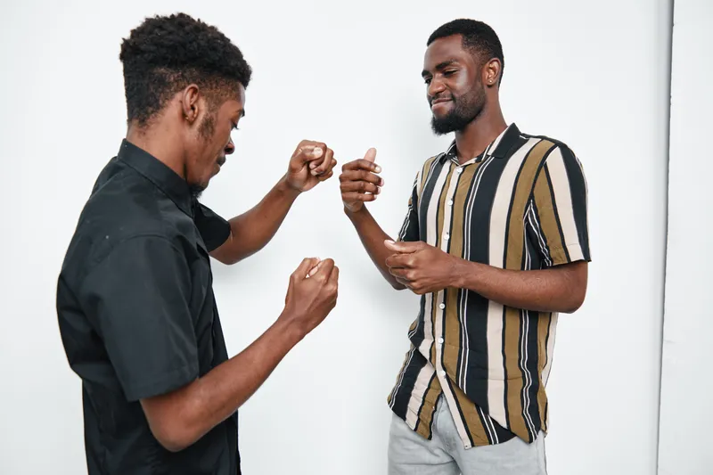 Two African American men with dark skin playing rock paper scissors game indoors. Casual clothing, friendly competition, and smiling expressions on white background.