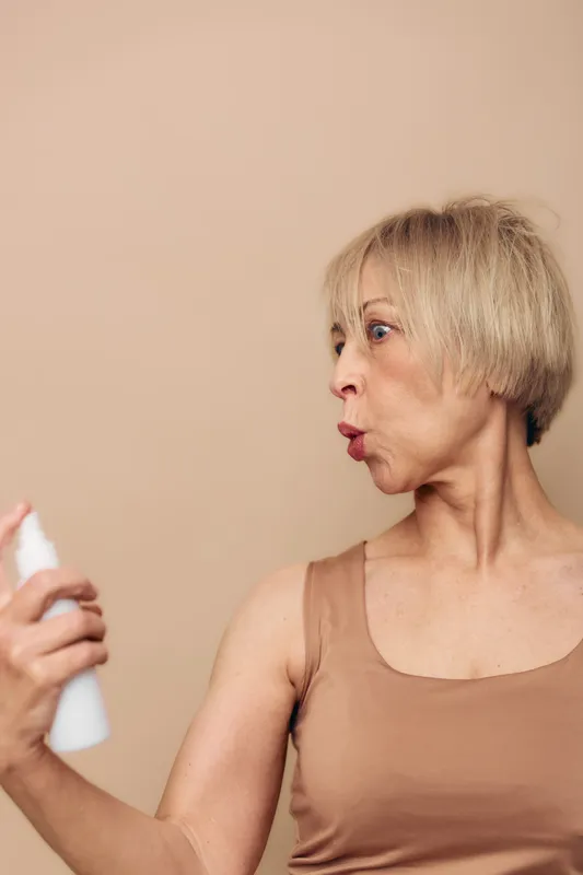 Middle aged woman with short blonde hair sprays facial mist on her face, wearing a sleeveless top. Concept of skincare, hydration, wellness, and self care in natural light neutral background.