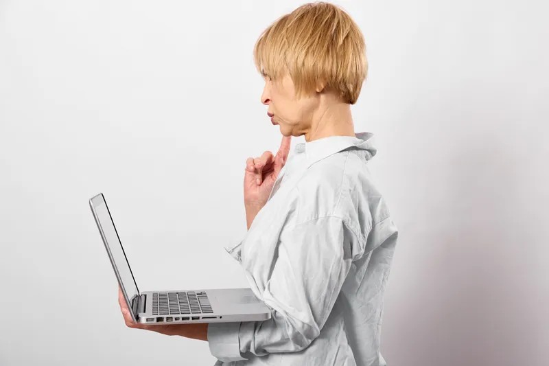 Stressed woman holding laptop close, hand on throat, showing pain and fatigue in workplace setup, side view on plain white background, concept for stress and health concern