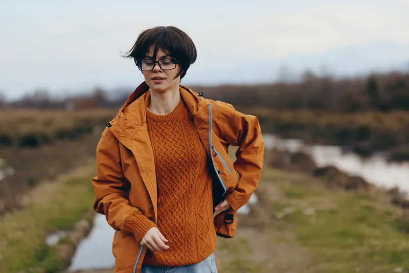 Woman outdoor lifestyle in autumn countryside, orange jacket and glasses, walking along rural path, relaxed expression, natural light, shallow depth