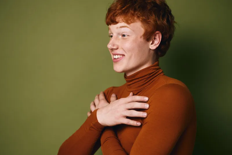 Young man with red hair smiling in brown turtleneck sweater crossing arms on green background in studio. Side view portrait with happy expression and casual style.