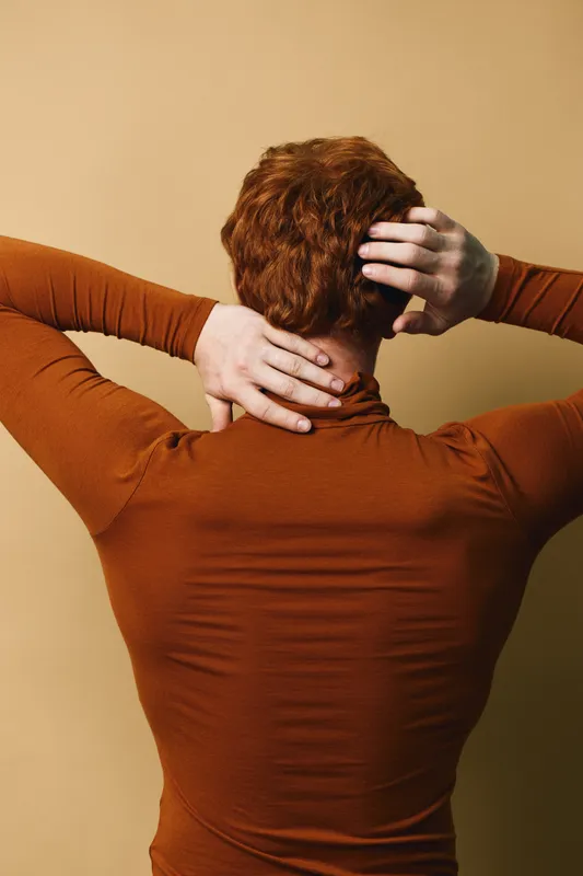 Person with red hair in brown long sleeve shirt standing with back to camera holding neck and head on beige background.
