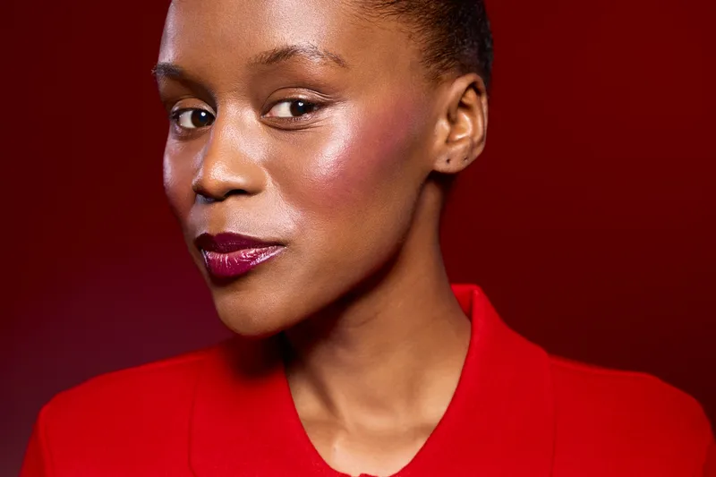 Confident African woman with dark skin and glowing makeup wears red outfit against dark red background. Portrait of beauty, fashion, and elegance with smooth skin and shiny lipstick.