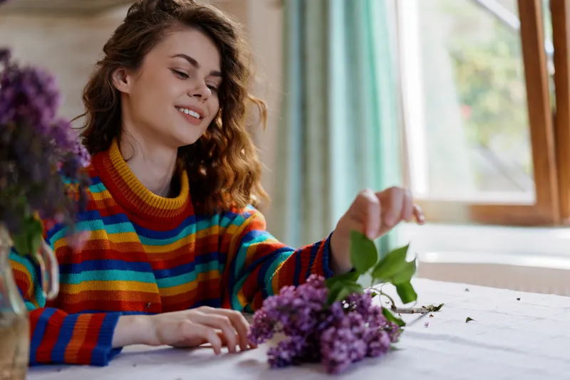 Young woman in a colorful striped sweater arranges a bouquet of lilacs on a sunlit table by a window, smiling softly as she enjoys a calm, intimate home moment.